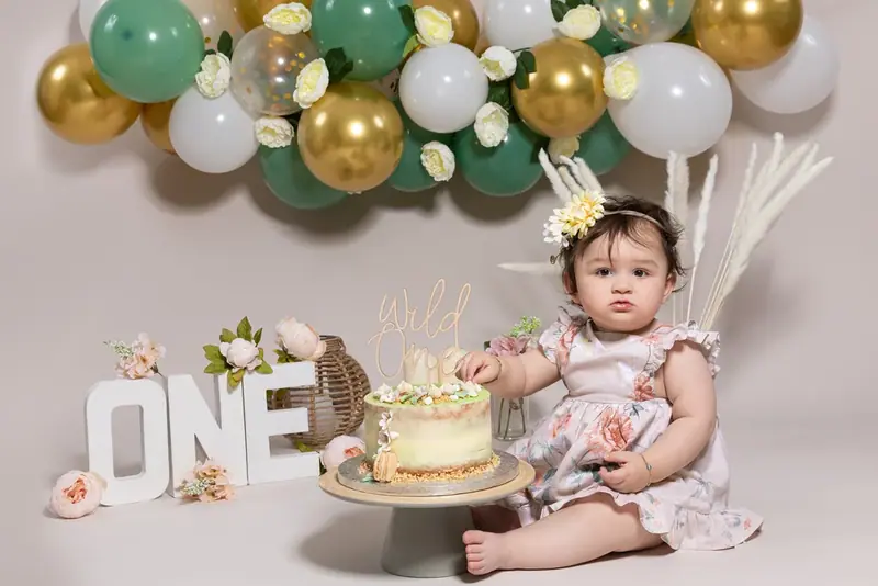 Baby beside a cake on a stand with green and gold balloons and the word ONE