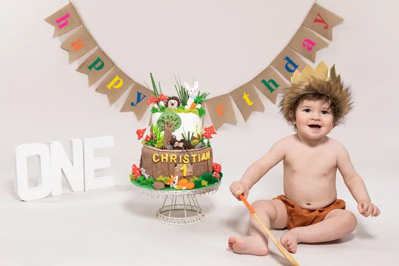 Baby in a crown beside a dinosaur birthday cake under a bunting banner