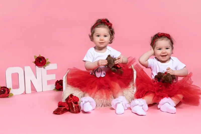 Two babies in red and white outfits sitting on a pink backdrop beside the word ONE