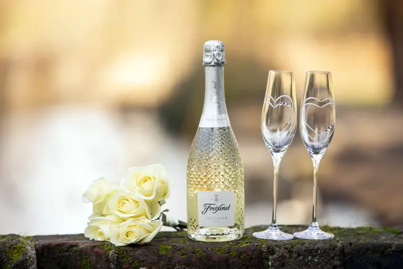 Bottle of sparkling wine with two glasses and pale roses on a wooden table