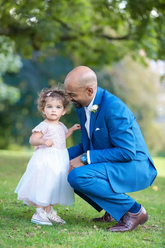 Groom in a blue suit crouching to talk to a small flower girl on the grass