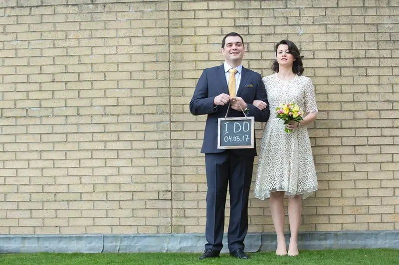 Bride and groom holding a framed sign in front of a pale brick wall