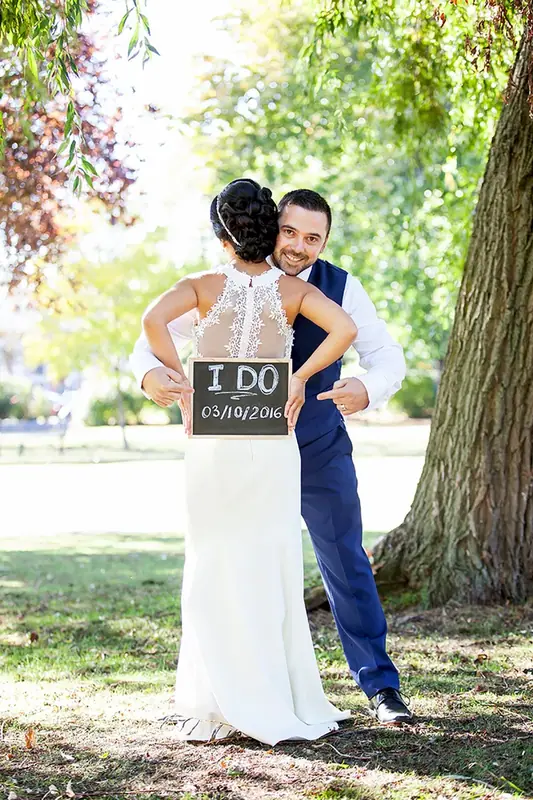 Bride and groom embracing beneath a tree, the groom holding a small sign