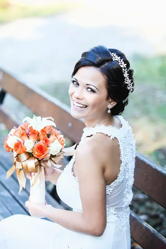 Bride in a lace dress holding an orange bouquet, seated on a bridge