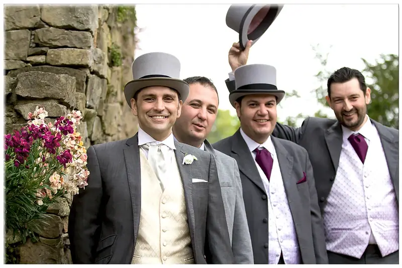 Groom and groomsmen in top hats posing beside a stone wall with flowers