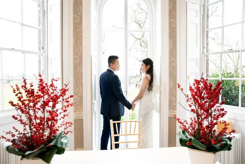 Bride and groom holding hands in a bright room decorated with red flowers