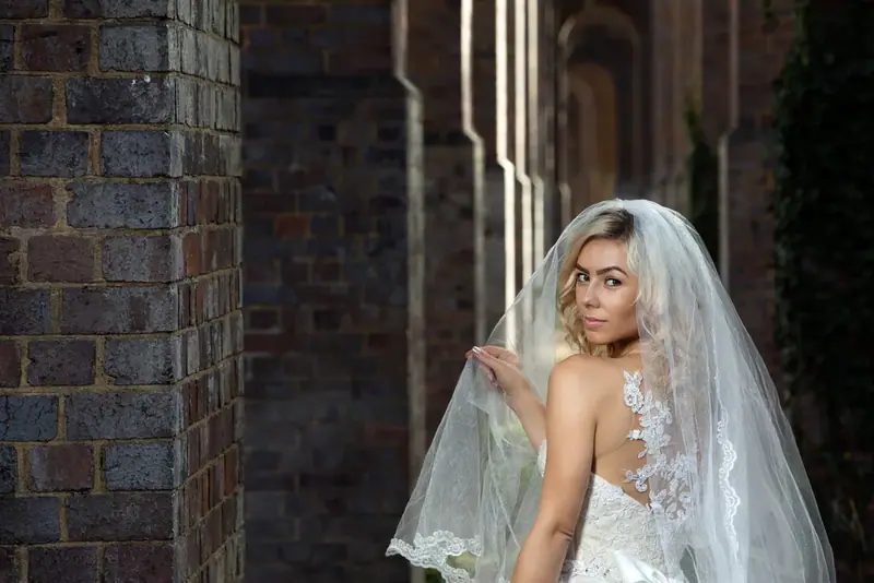 Bride adjusting her veil beside an ivy-covered brick column