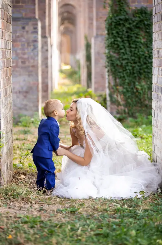 Bride crouching to hug a small boy in a blue suit among brick arches
