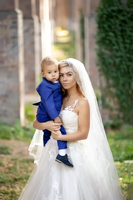 Bride holding a small boy in a blue suit in front of ivy-covered arches