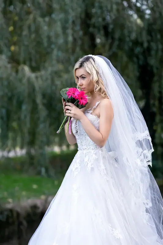 Bride in a veil holding a red bouquet under a willow tree
