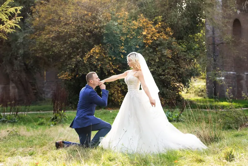 Groom kneeling to kiss the bride's hand on a lawn in front of an old building