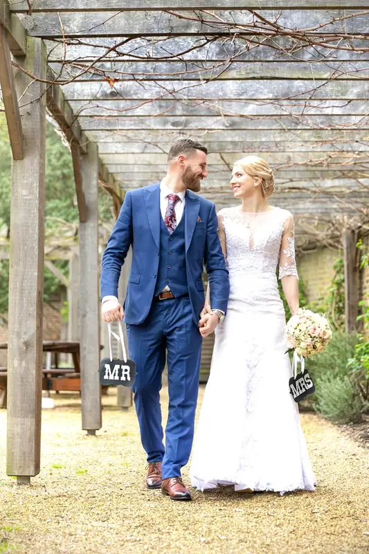Bride and groom in a blue suit walking beneath a wooden pergola