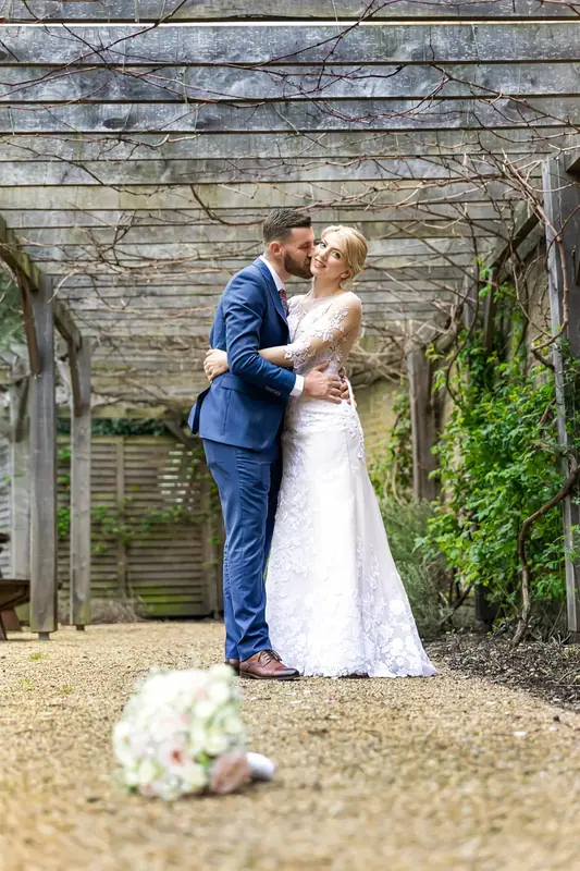 Bride and groom embracing beneath a wooden pergola with a bouquet